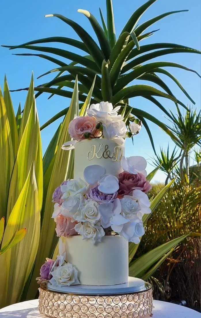 A white wedding cake adorned with pink, purple, and white flowers, placed outdoors against tall green plants and a blue sky.