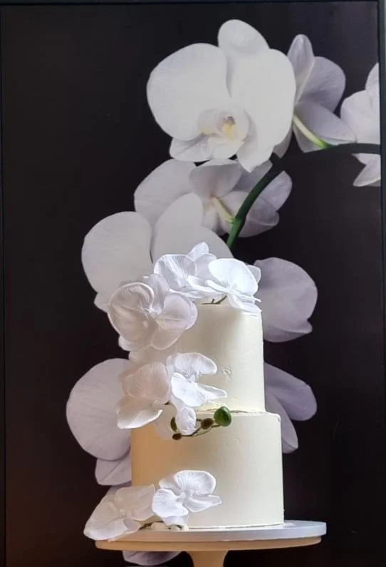 A two-tiered white cake decorated with white orchids in front of a large floral backdrop.