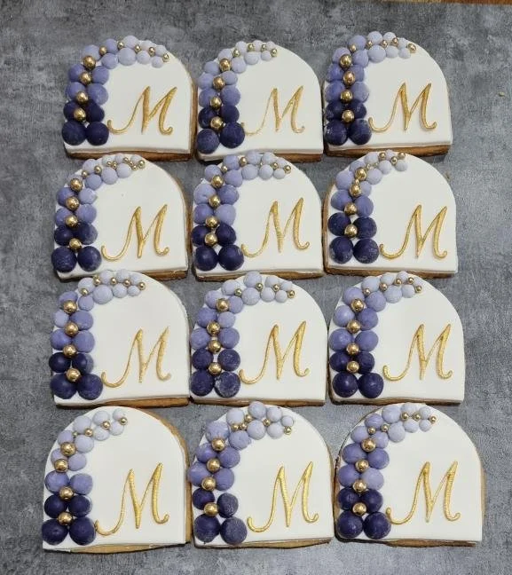 Eleven decorated cookies on a gray surface, each with a white icing base, a gold letter 'M' in the center, and a pattern of purple, lavender, gold, and white spherical decorations on one side.