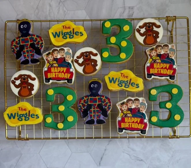 Decorative cookies on a wire rack celebrating a child's third birthday with themes from The Wiggles and characters from a children’s show, including large number 3 cookies, The Wiggles logo cookies, and character cookies.