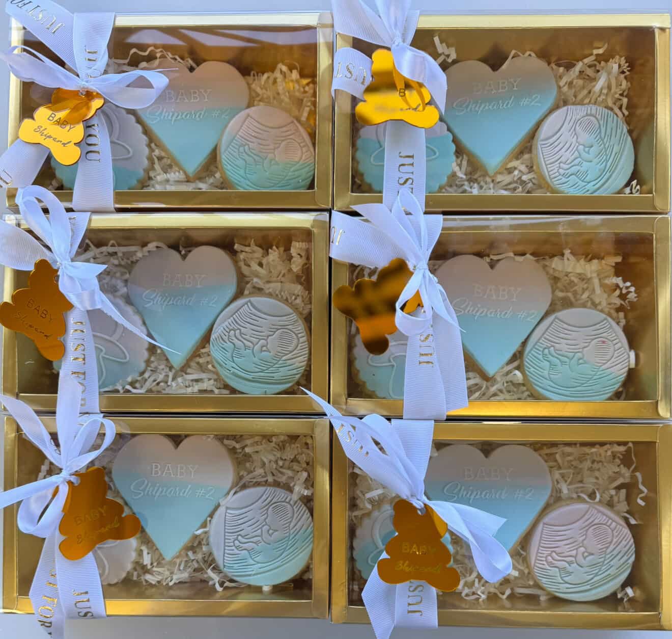 Six gift boxes with clear windows containing decorated cookies in the shape of hearts, circles, and baby footprints, labeled 'Baby Shrimp #2' with white ribbons and small gold tags.