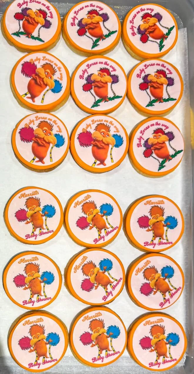 Cookies with cartoon characters and phrases for a baby shower, featuring a fluffy bird holding colorful pom-poms and the phrase "Baby Love on the way" and a bird with pom-poms with the text "Harriet Baby Shower."