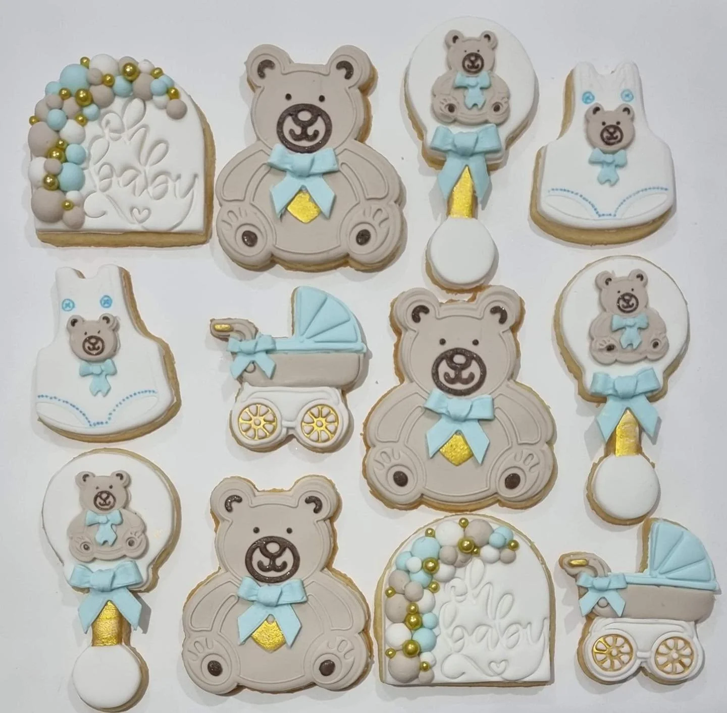 Assorted decorated cookies with teddy bear, baby stroller, bib, and Easter egg designs in pastel colors with blue, white, gold, and beige accents.