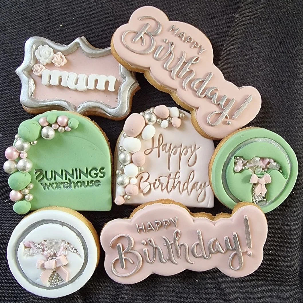 Collection of decorated birthday cookies with messages like "Happy Birthday," "mum," and "Bunnings Warehouse," and designs including pastel pink, white, and green icing with silver and pink sprinkles, some with floral or bow accents.
