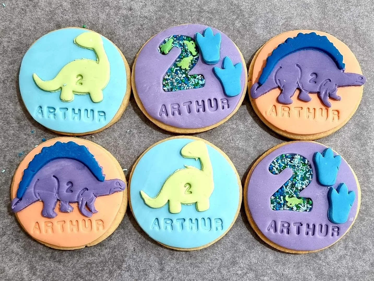 Set of six colorful decorated cookies on a gray surface. Each cookie features a dinosaur silhouette with the number 2 and the name 'Arthur' in different pastel icing colors, some with decorated sprinkles.