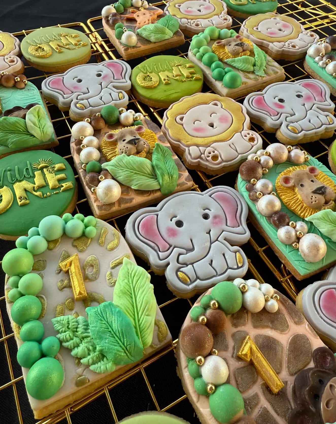 Decorated cookies with elephants, lions, and other animals, featuring green, white, gold, and neutral colors, arranged on a wire cooling rack.