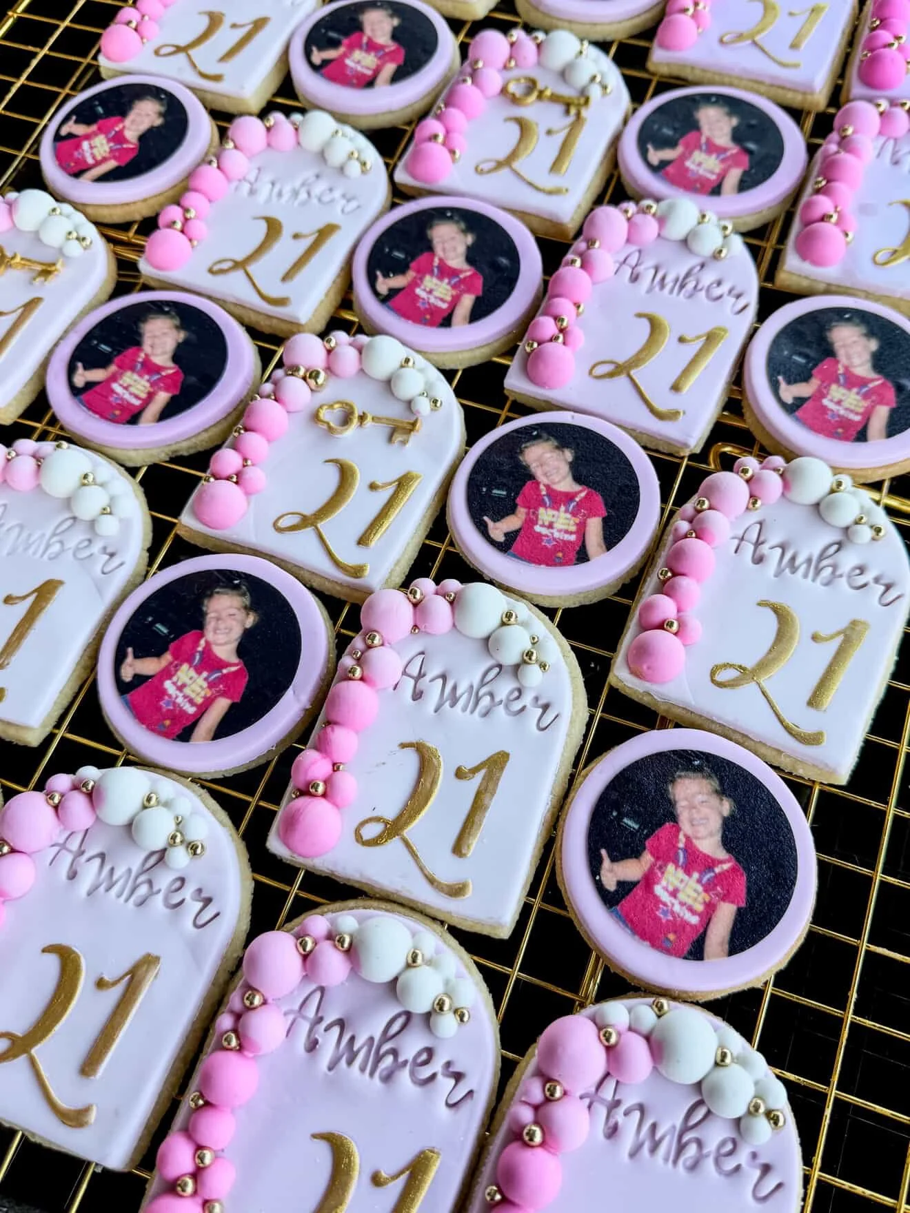 Decorated cookies celebrating a 21st birthday, with some featuring a young girl giving a thumbs up, and others with the words 'Happy 21st' written on them, along with pink and white bead decorations.