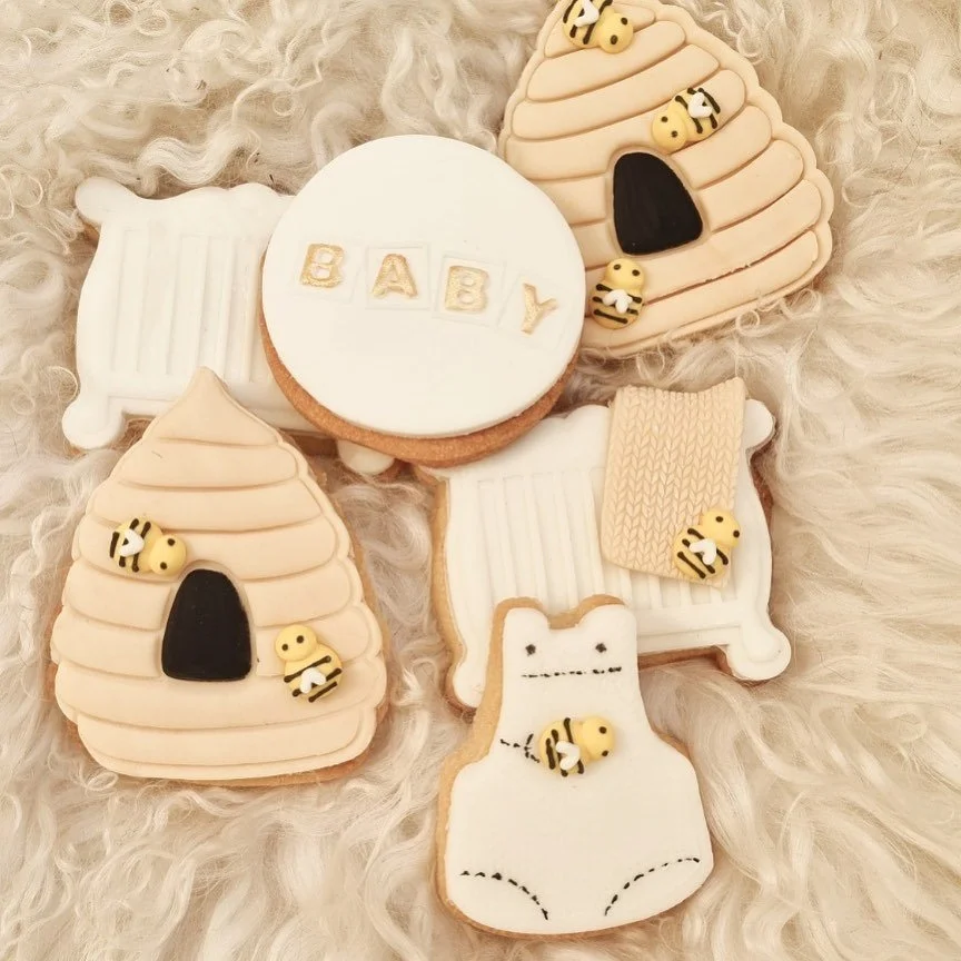 Decorated cookies with baby-themed designs on a fluffy surface. Designs include bumblebees, a white bear, baby cribs, a honey jar, and a cookie with the word "BABY" written on it.