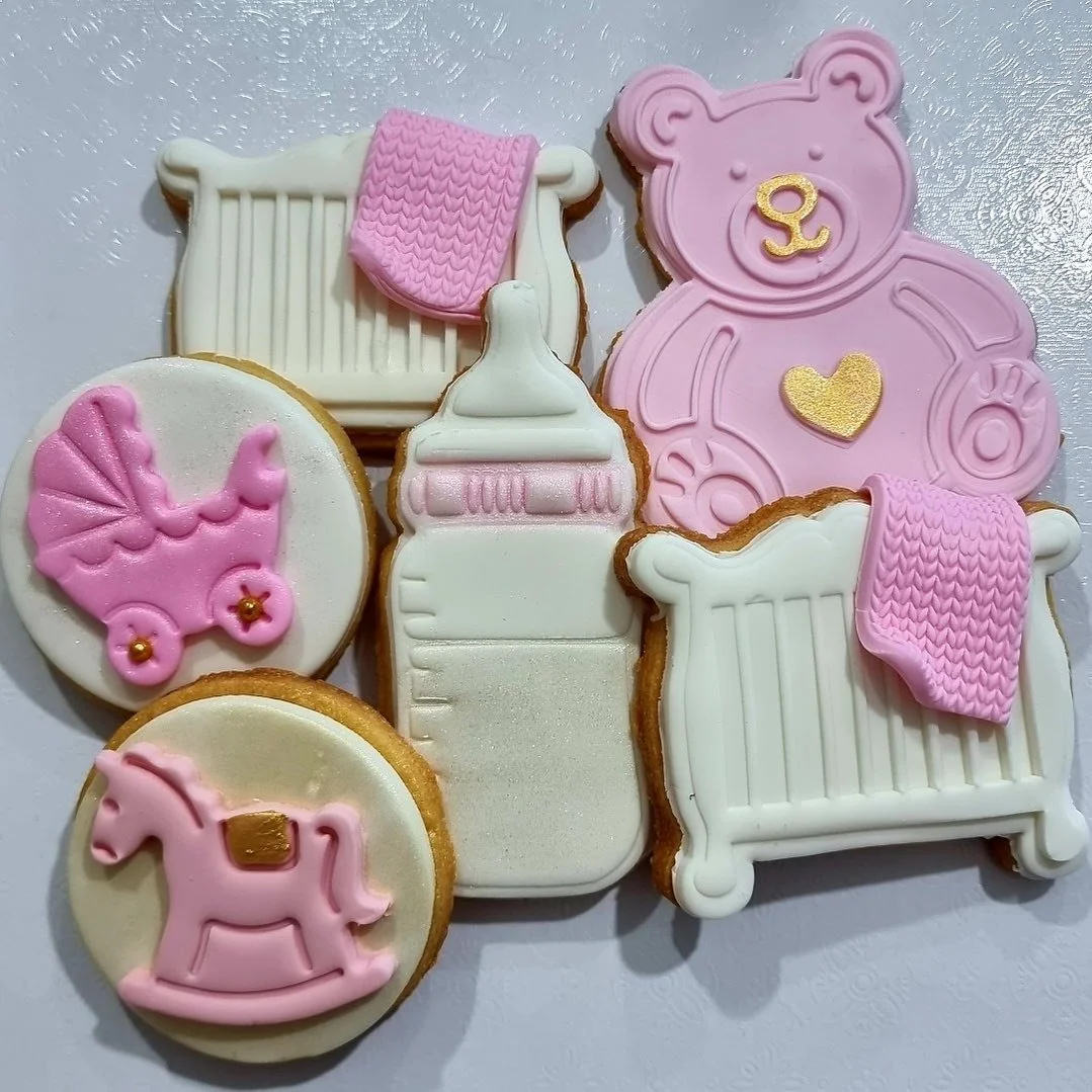 Decorated cookies with baby and nursery themes, including a teddy bear, bottles, a cradle, a rocking horse, and a teddy bear face, in pink, white, and gold colors.