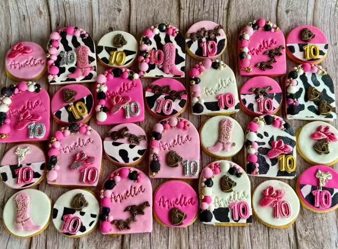 Assorted decorated cookies arranged on a wooden surface, celebrating a 10th birthday for someone named Amelia. The cookies feature pink, black, and white colors with various designs including hearts, cow patterns, and the number 10.