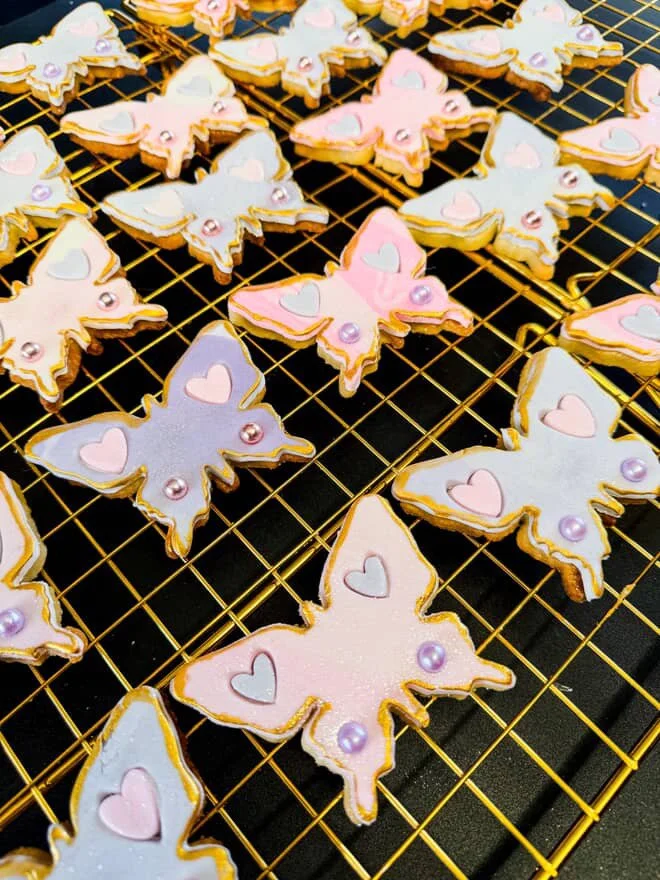 Decorated butterfly-shaped cookies with pastel pink, purple, and blue icing, adorned with small heart-shaped and round sugar decorations, cooling on a wire rack.