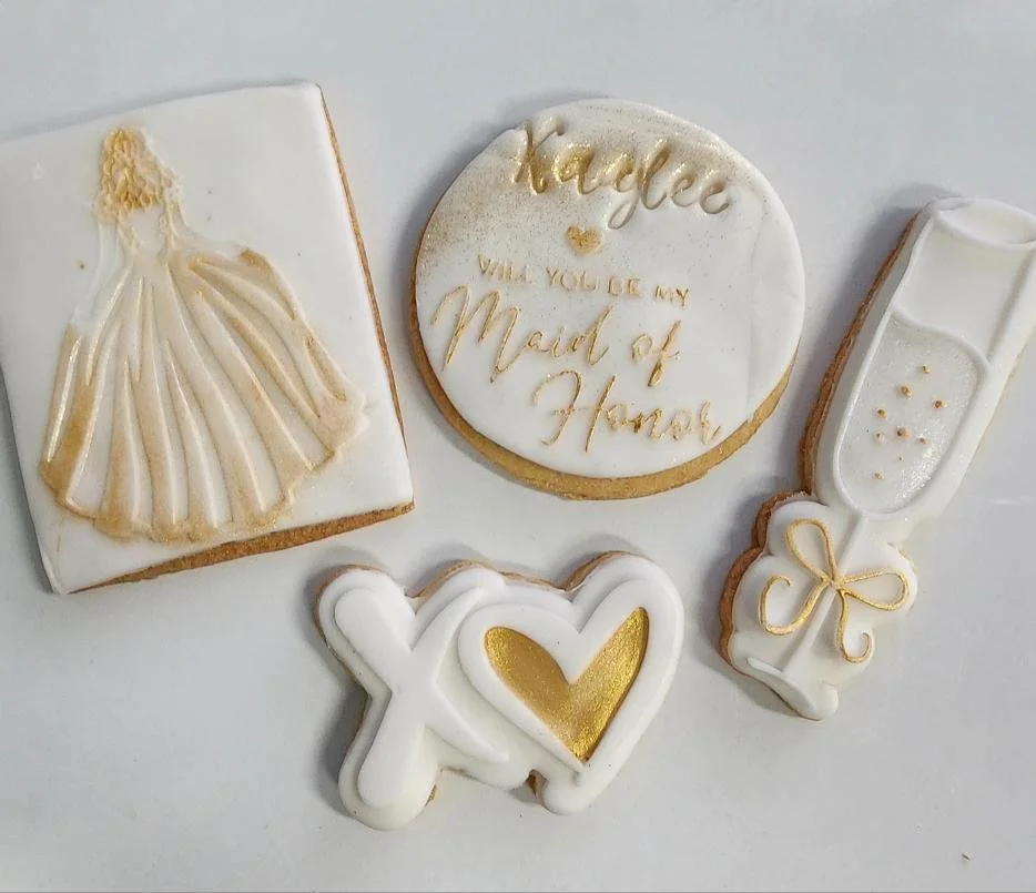 Cookies decorated with white and gold icing, including a wedding dress, a champagne glass with a bow, a heart with intertwined letters, and a round cookie with the message 'Hooray! Will you be my Maid of Honor?'