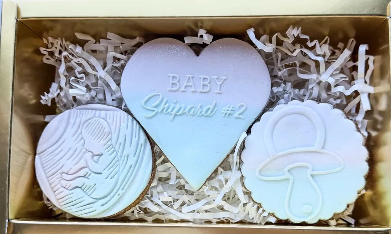 A gift box with three decorated cookies: one shaped like a heart with the words "BABY Shipord #2", one with an outline of a baby's face, and one featuring a mushroom with a simple design.