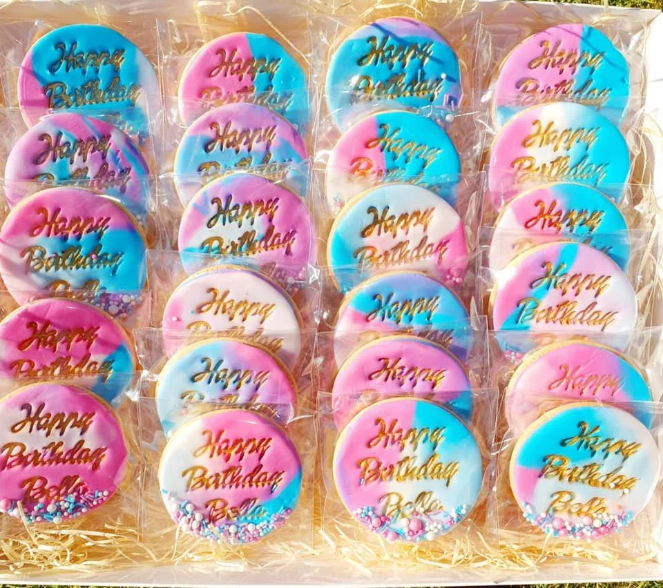Decorated cookies with pastel icing, each inscribed with 'Happy Birthday' in gold lettering, enclosed in clear plastic wrapping, arranged in rows.