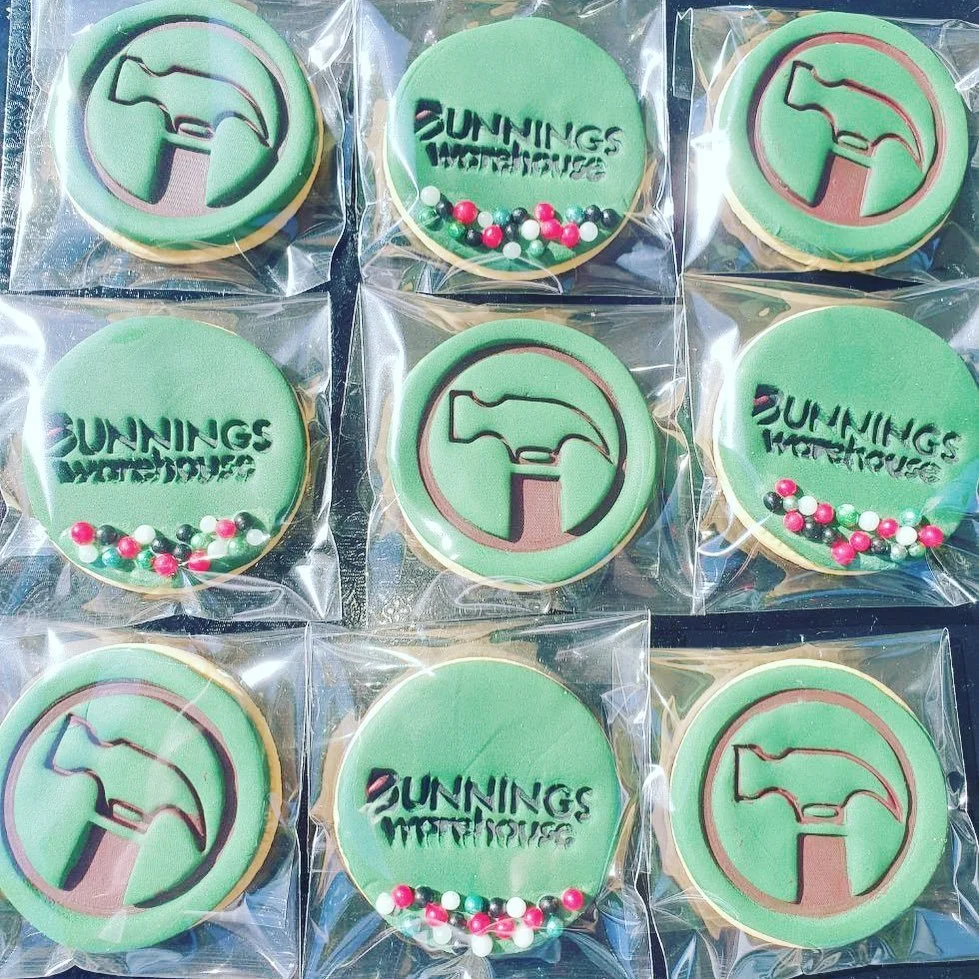 Assorted cookies with green icing in plastic wrappers, featuring a hammer logo and the text 'SUNINGS WEATHER DICES' with decorative pink, white, and black sugar beads at the bottom.