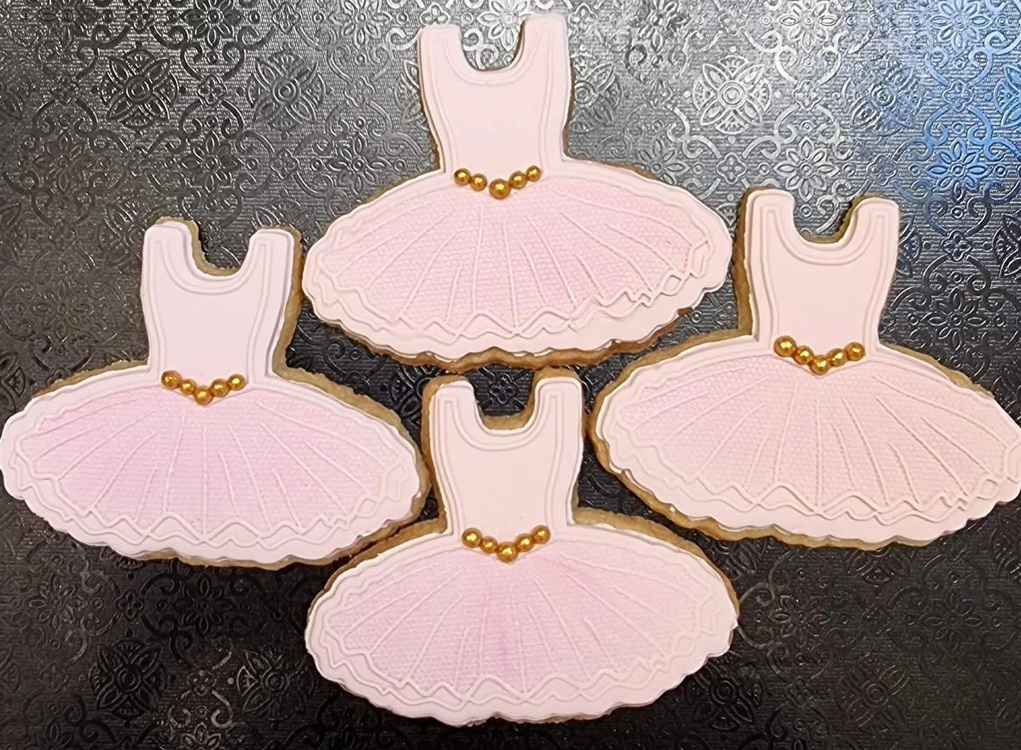 Ballet slipper-shaped cookies with pink icing and gold bead decoration, arranged on a black patterned surface.