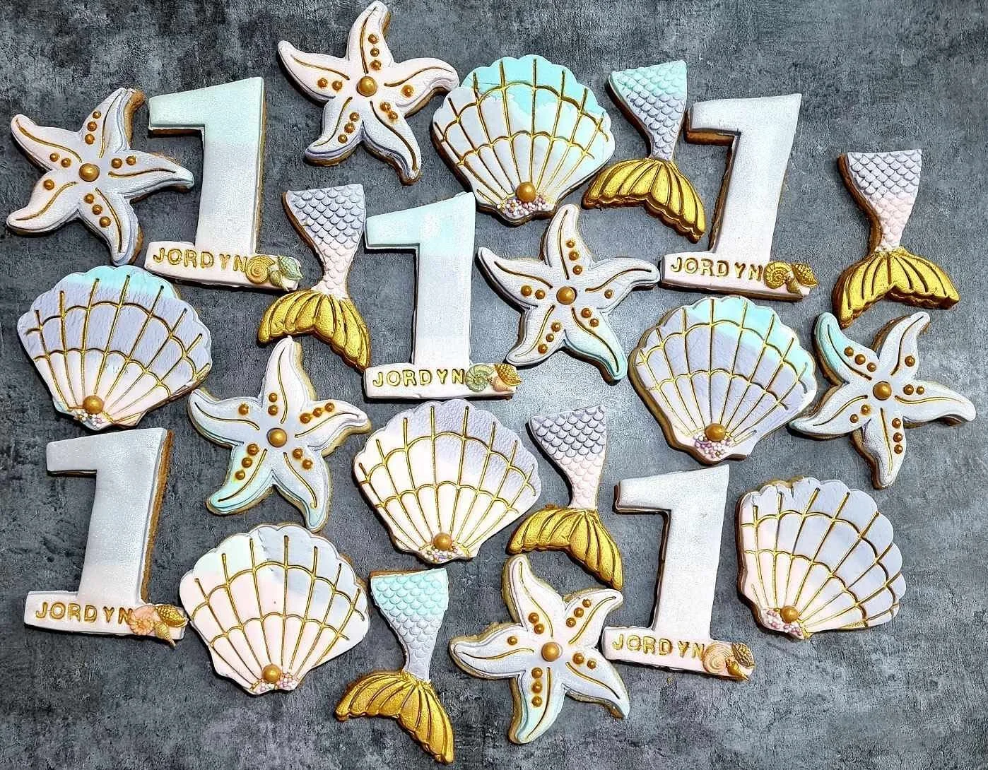 Decorative cookies shaped like the numbers 1, 2, and 3, with seashell and starfish designs, arranged on a gray surface. Some cookies are inscribed with the name 'JORDYN'.