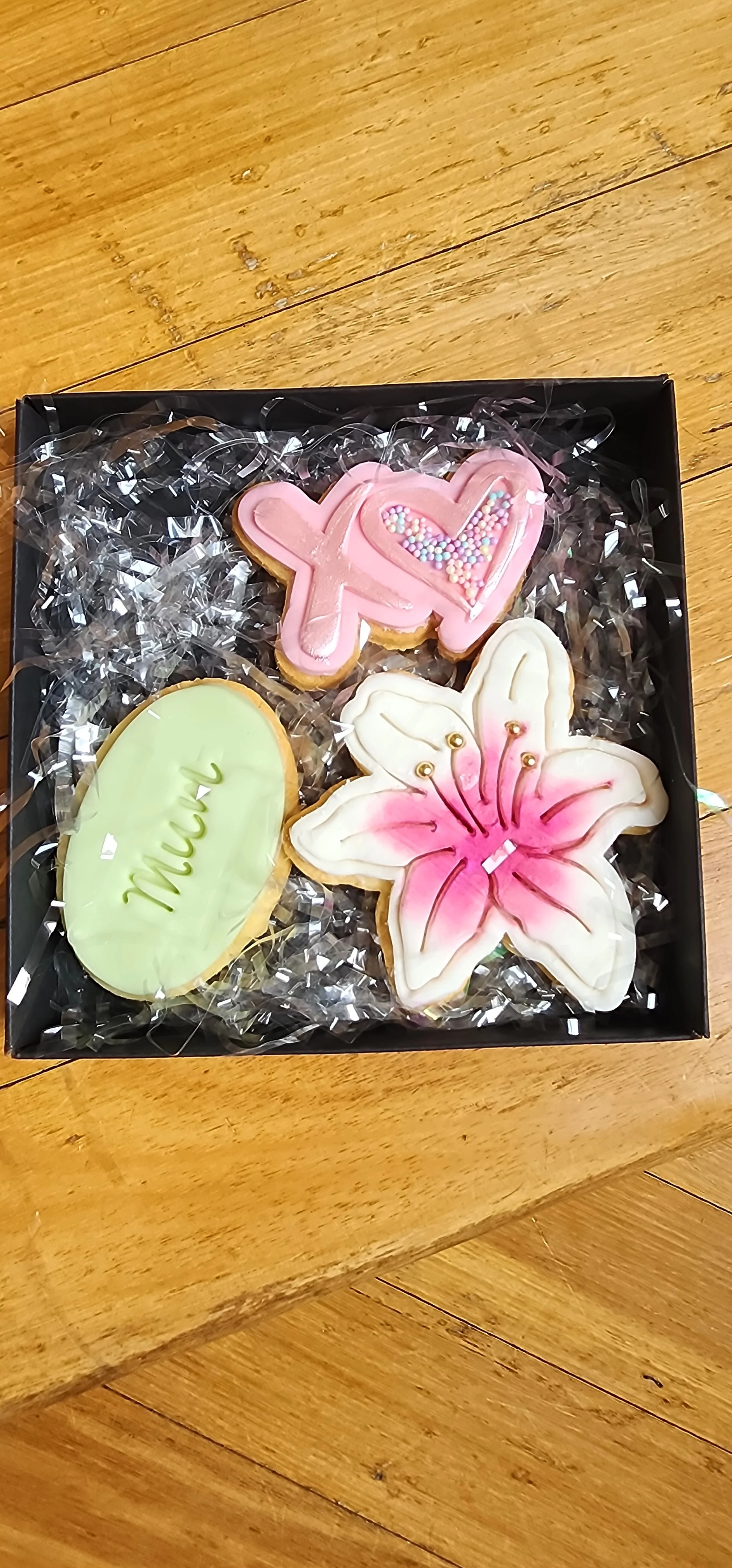 A black box containing three decorated cookies on a bed of clear curly plastic wrap. One cookie is shaped like a pink hello signal with heart and rainbow sprinkles, another is an oval with light green icing and the word 'mum' written on it, and the t