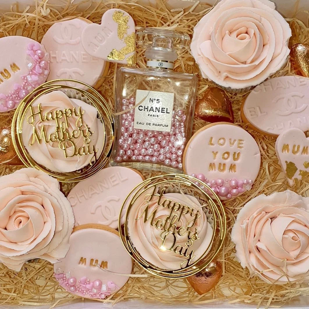 A Mother's Day gift box containing pink roses, decorated cookies with messages like "Happy Mother's Day" and "Love You Mum," a Chanel perfume bottle, pink pearl beads, and gold heart-shaped chocolates, arranged on shredded paper.