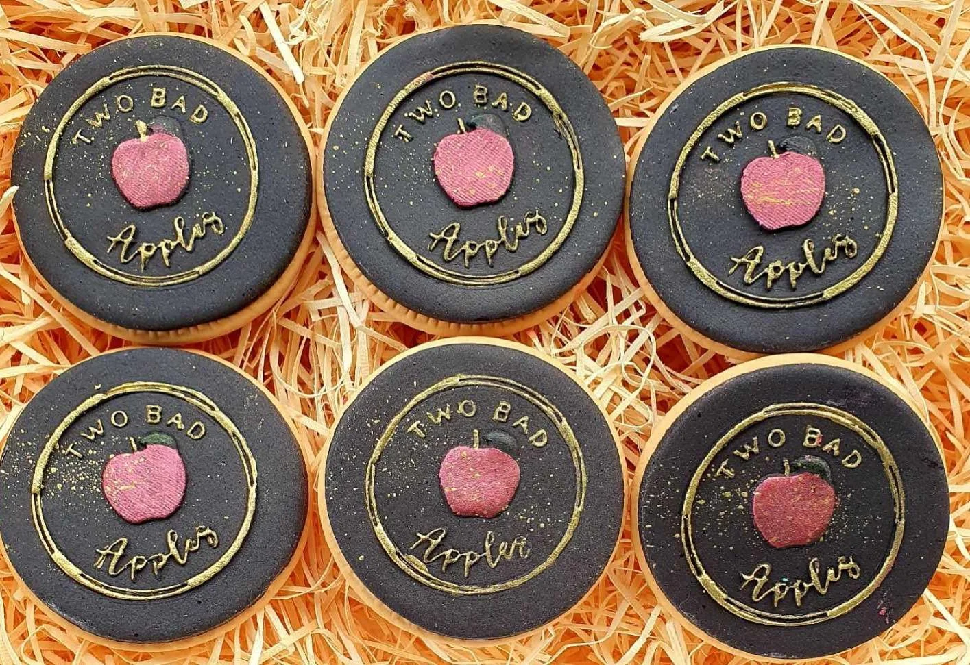 Six round cookies with black icing, each decorated with an apple and the phrase "TWO BAD Apple" in gold lettering, resting on shredded orange paper.