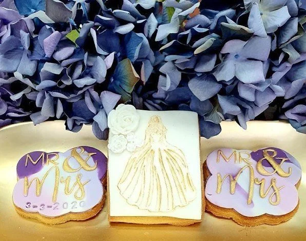 Wedding cookies with purple and gold icing spelling 'Mr. & Mrs.' and a decorated bride silhouette, placed on a gold tray with blue hydrangea flowers in the background.