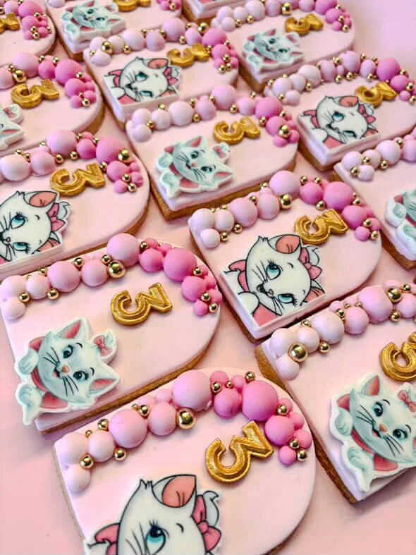 Decorative cookies with pink icing, featuring cartoon kittens, pink and gold beads, and a gold number 3, arranged on a pink surface.