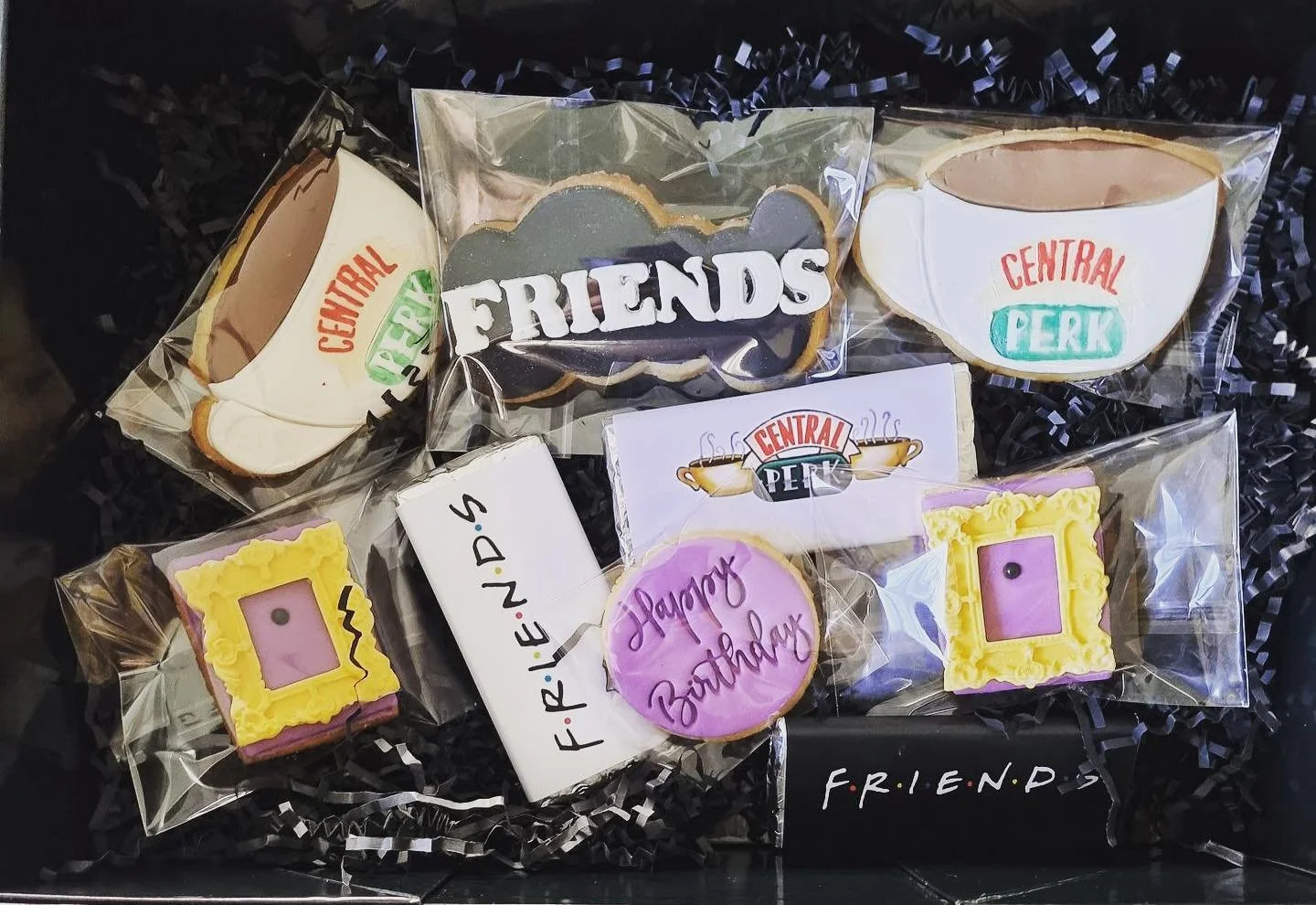 A collection of 'Friends'-themed cookies and chocolate treats, including cookies decorated as Central Perk coffee cups, tasteful cookies with 'FRIENDS' and 'Happy Birthday' messages, and others with yellow and purple frames, all stacked in a box with