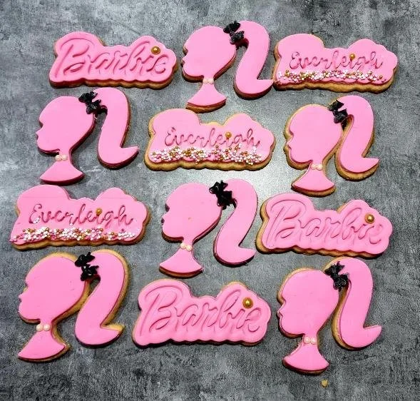 Assorted Barbie-themed cookies in pink, shaped as Barbie's profile, the word 'Barbie,' and 'Eureka!' with decorative sprinkles, arranged on a gray surface.