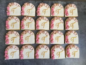 Sixteen decorated cookies arranged in four rows, with a design that includes pink and white icing, pearls, and a gold letter 'T' in the center.