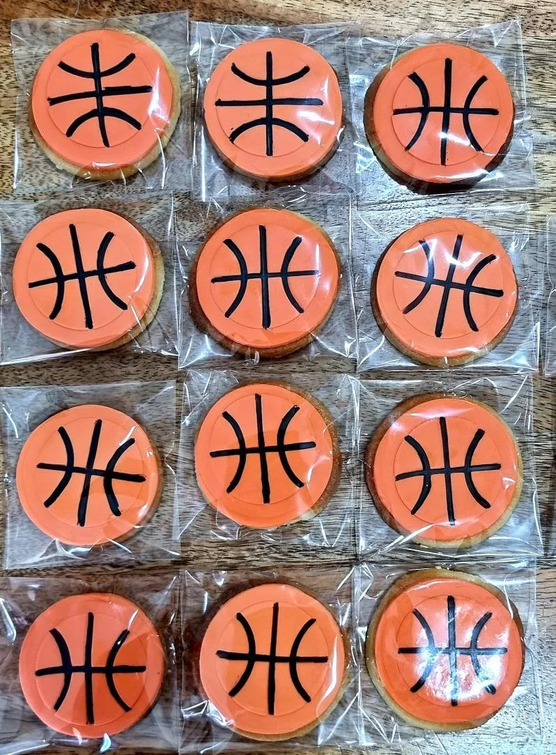 Cookies decorated with orange icing and a black basketball emblem, individually wrapped in clear plastic, arranged on a wooden surface.