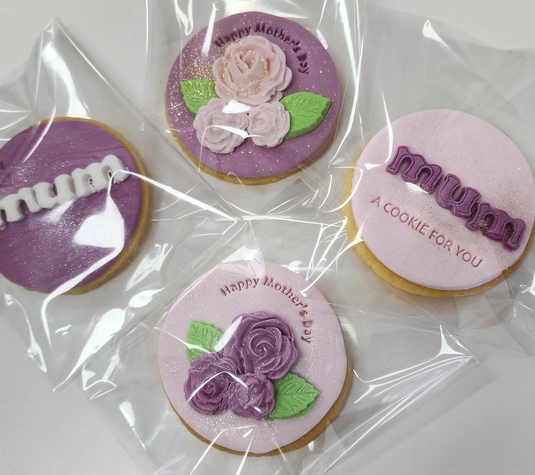 Decorative cookies with floral and message designs, wrapped in clear plastic. One cookie has a purple background with pink roses and green leaves, with "Happy Mother's Day" written. Another cookie is pink with the word "Mom" in purple. A third cookie