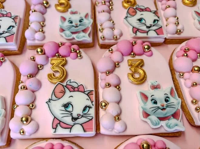 Pink cookies decorated with cartoon cat images, pink and gold beads, and a gold number three for birthday celebration.