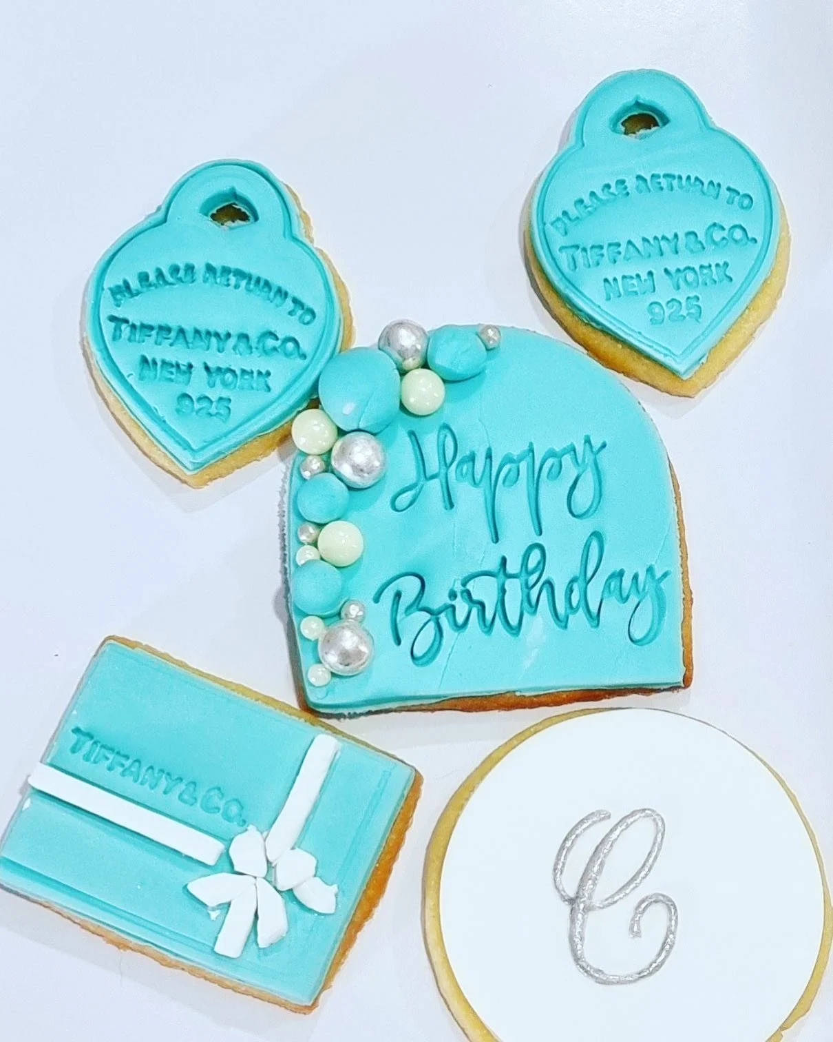 Set of Tiffany blue-themed cookies for a birthday celebration. Includes a large 'Happy Birthday' cookie decorated with pearls and small blue candies, two heart-shaped cookies with Tiffany & Co. branding, a square cookie resembling a Tiffany box with 
