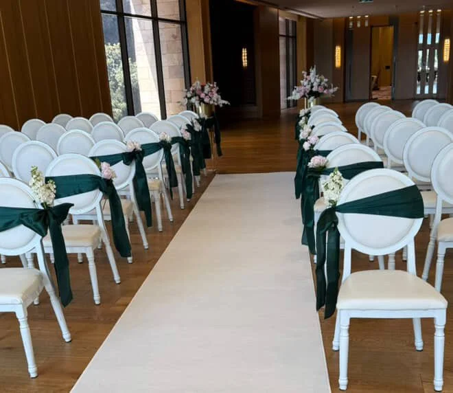 Wedding ceremony chairs arranged on either side of an aisle in a decorated room with flowers and draped fabric.
