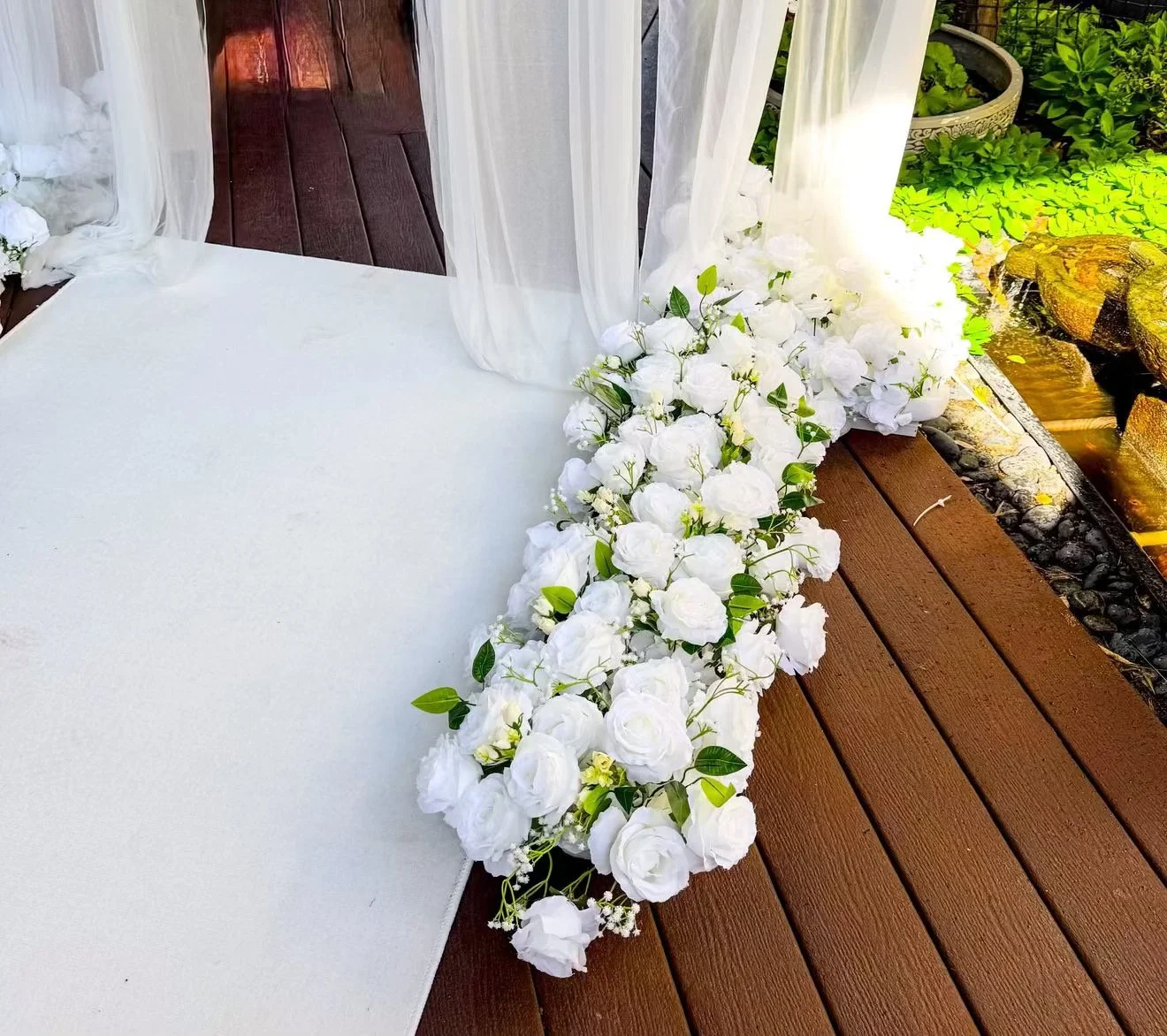White floral arrangement with roses and greenery on a wooden deck near a small garden with water feature.