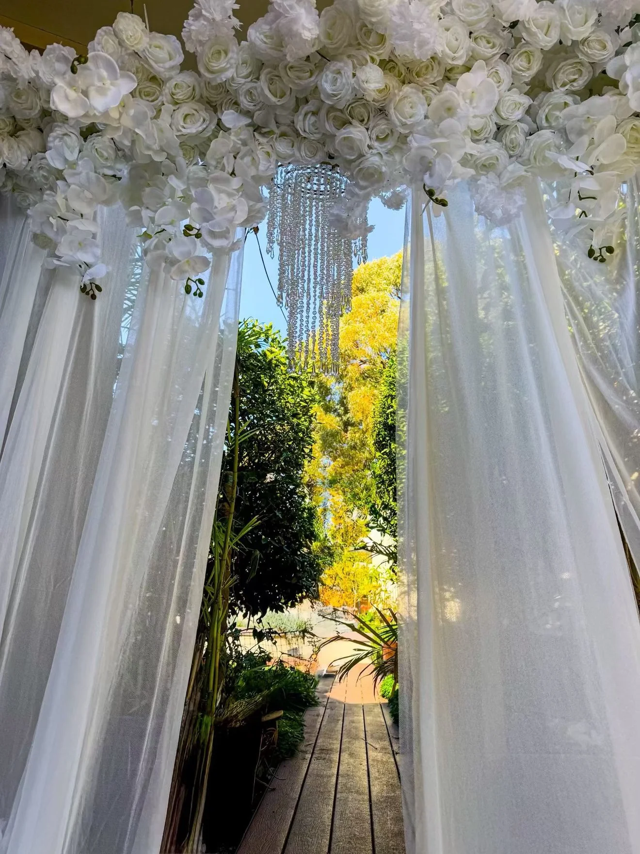 Flowing white curtains framing a garden pathway with lush green and yellow foliage, a chandelier hanging above, and a clear blue sky in the background.