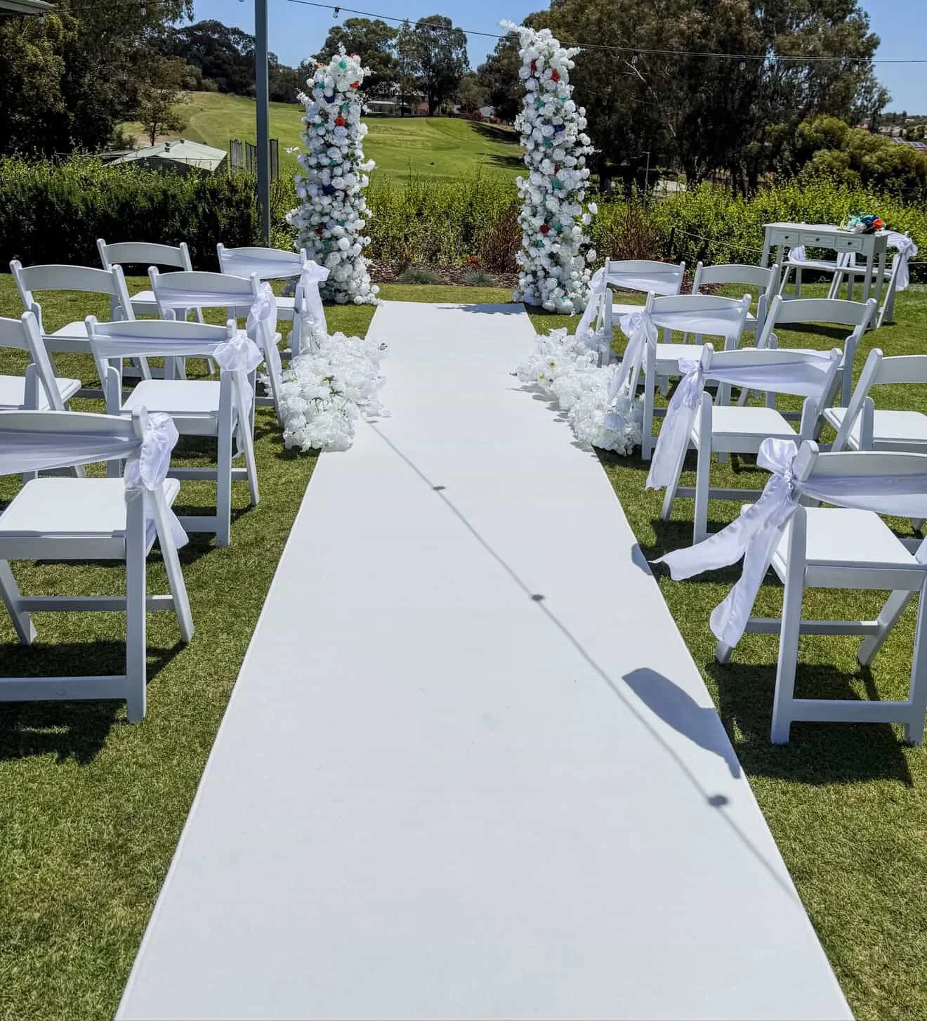 Outdoor wedding ceremony setup with white chairs decorated with white ribbons arranged on both sides of an aisle, leading to a floral arch with white and colored flowers, set in a grassy area with trees and a hill in the background.