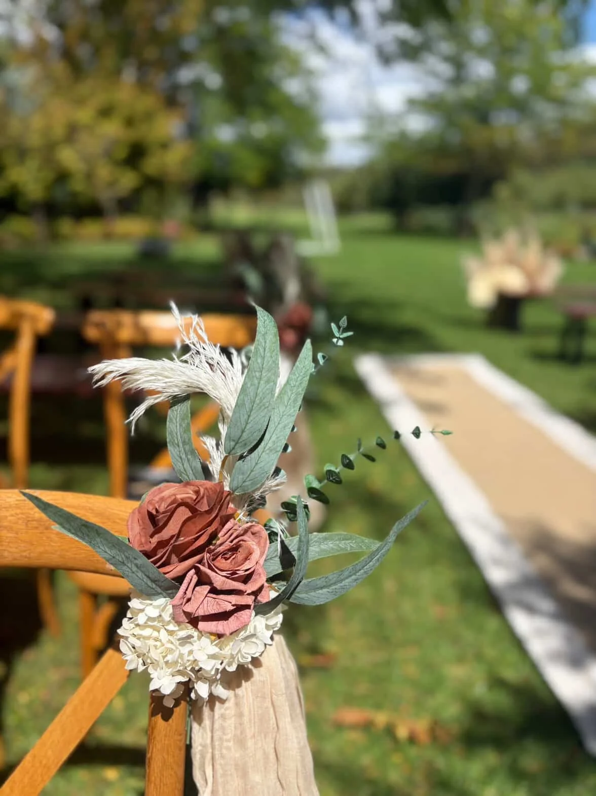 Close-up of a flower arrangement attached to a wooden chair at an outdoor event, with a grassy garden and pathway in the background.