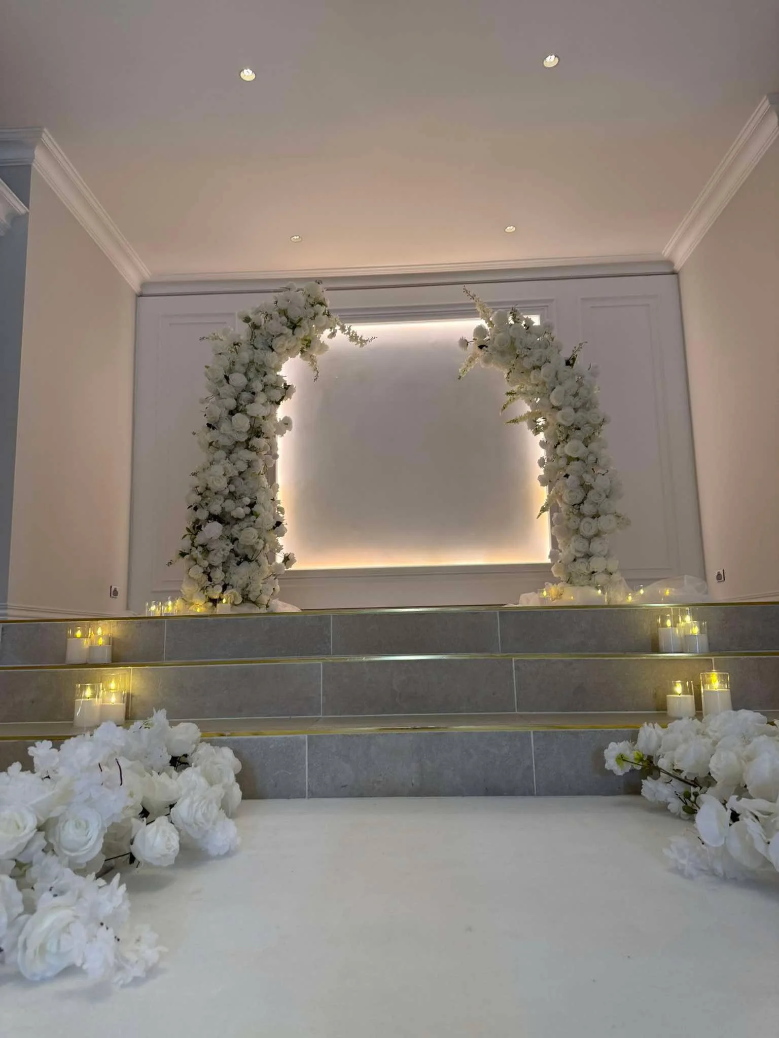Wedding altar decorated with white flowers, illuminated backdrop, surrounded by white floral arrangements and candles, in a modern indoor setting.