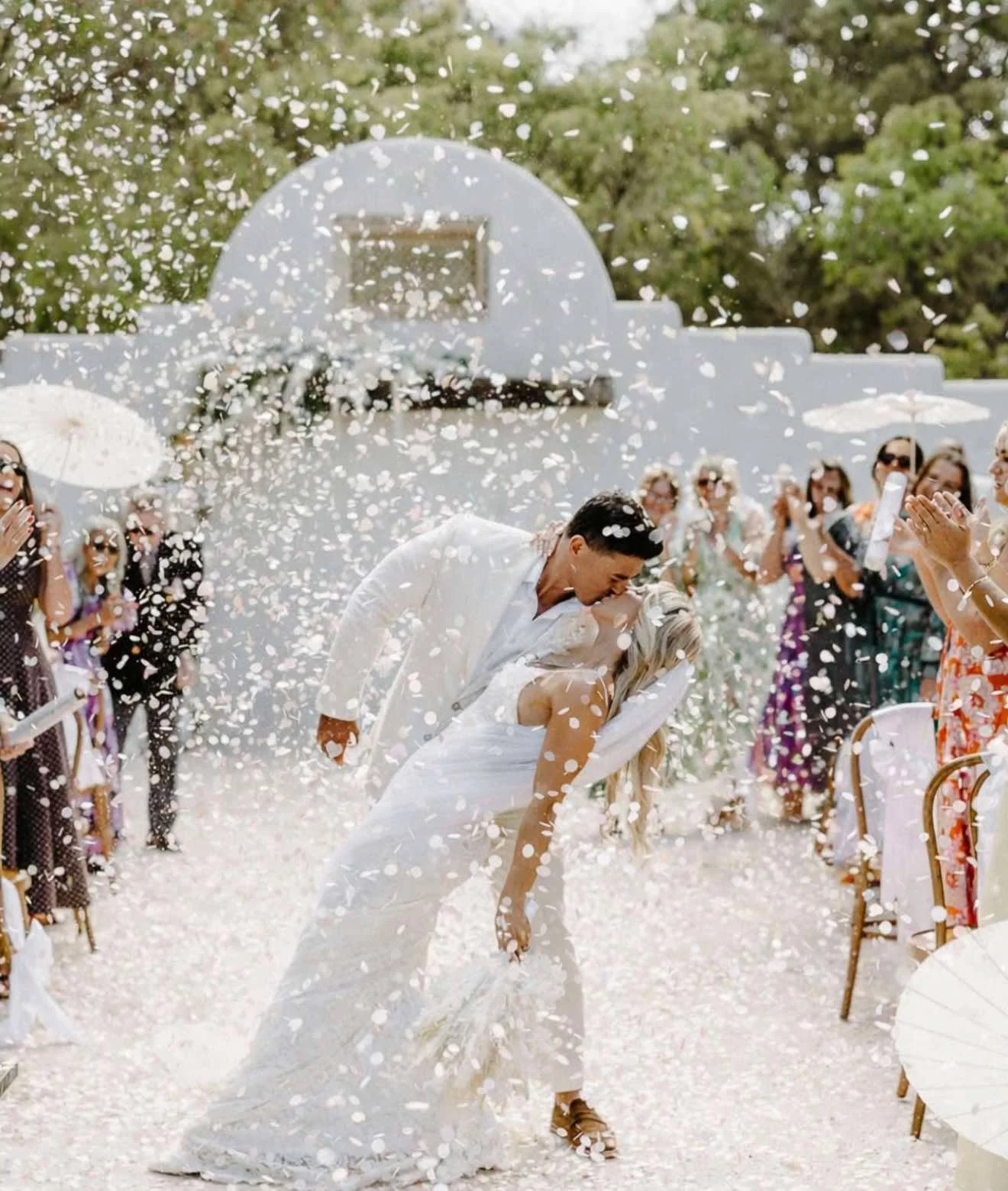 A bride and groom share a kiss while popping champagne at their outdoor wedding ceremony, with guests applauding and celebrating around them.