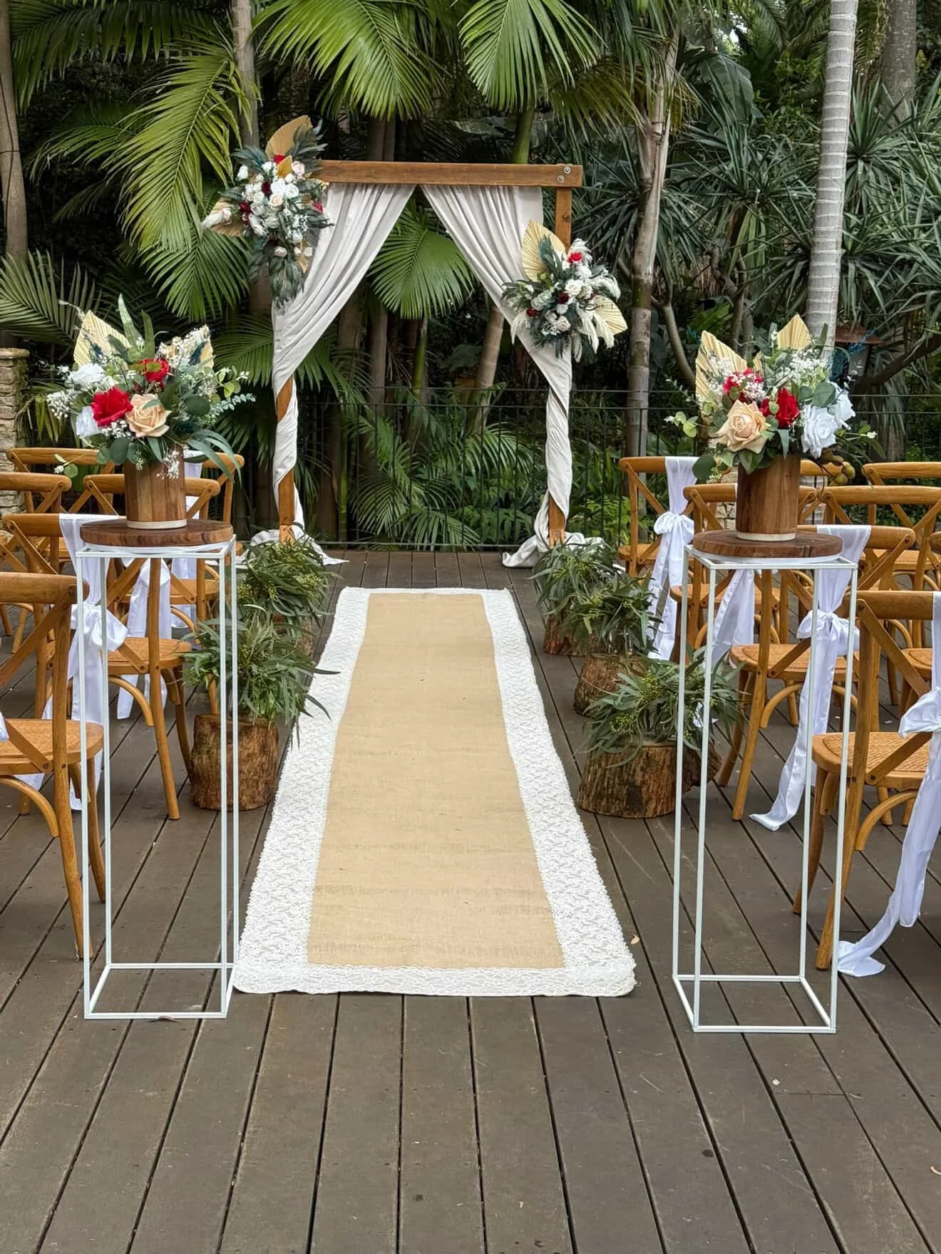 A beautifully decorated outdoor wedding altar with a wooden arch, white curtains, floral arrangements, and a beige and white runner on a wooden deck surrounded by lush greenery.
