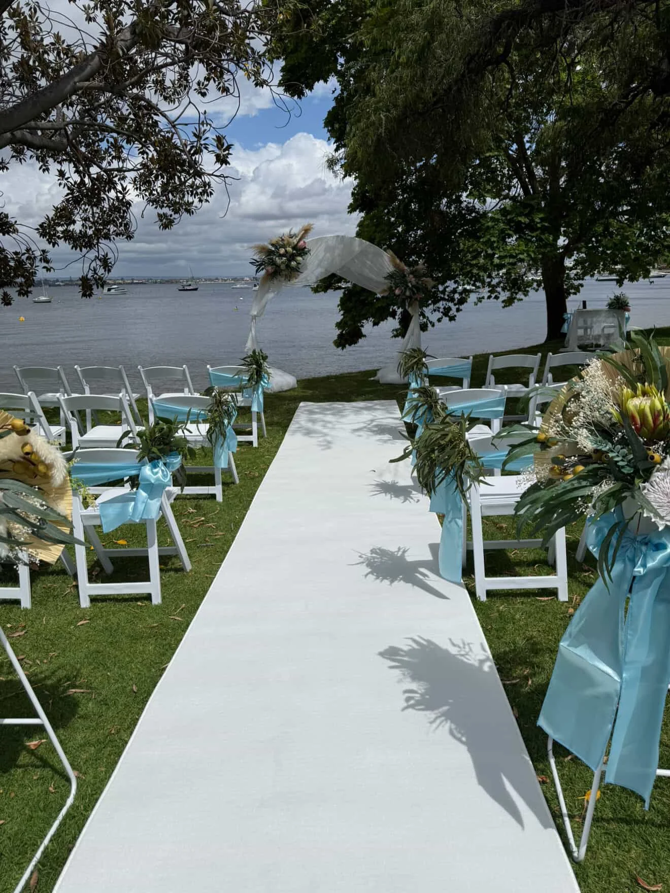 Outdoor wedding ceremony setup by a lake with white chairs decorated with blue ribbons, a white aisle runner, floral arrangements, and a decorative arch with flowers under trees with a view of water and boats in the background.