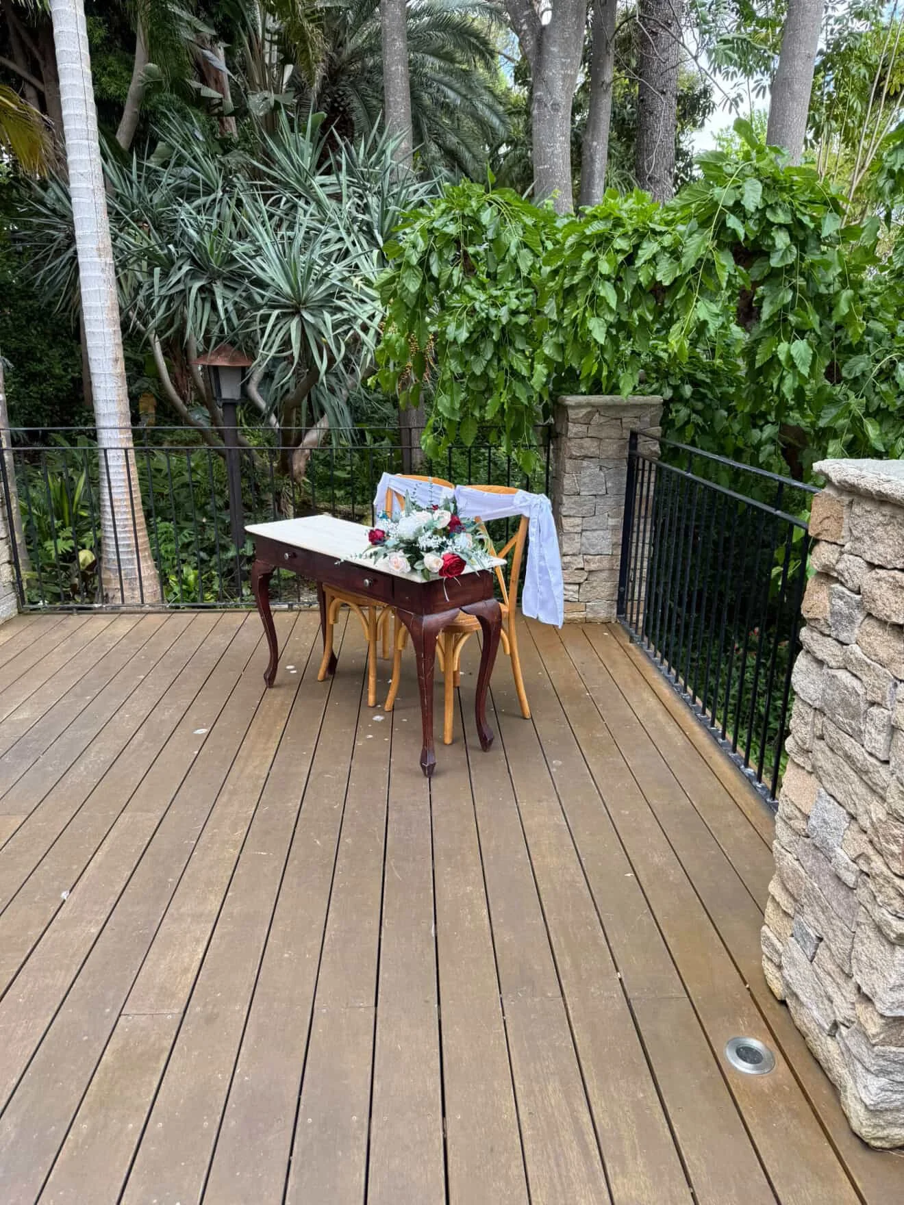 An outdoor patio with wooden decking, a small table decorated with a floral arrangement and a white cloth, surrounded by trees and lush greenery, with stone and metal fencing.
