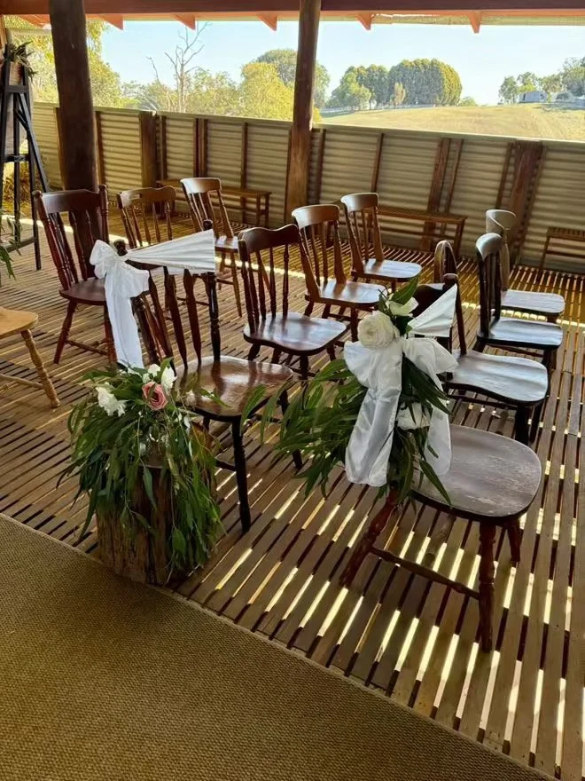 Chairs decorated with white ribbons and flowers, arranged in a semi-circle on a wooden deck, with a large flower arrangement in the foreground.