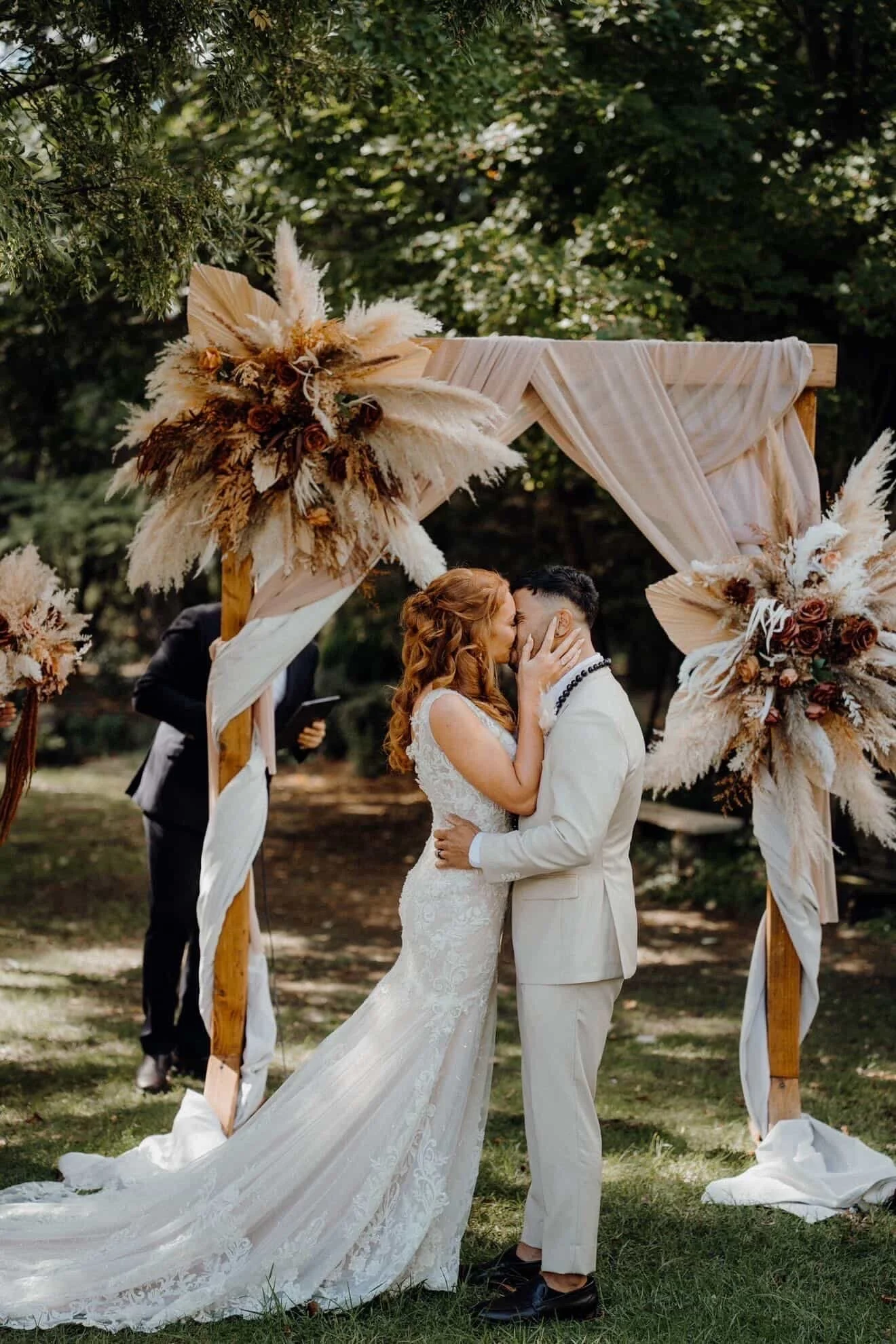 A bride and groom sharing a kiss during their outdoor wedding ceremony beneath a decorated wooden arch with dried floral arrangements. The bride wears a white lace gown with a long train, and the groom is in a light-colored suit. The ceremony takes p