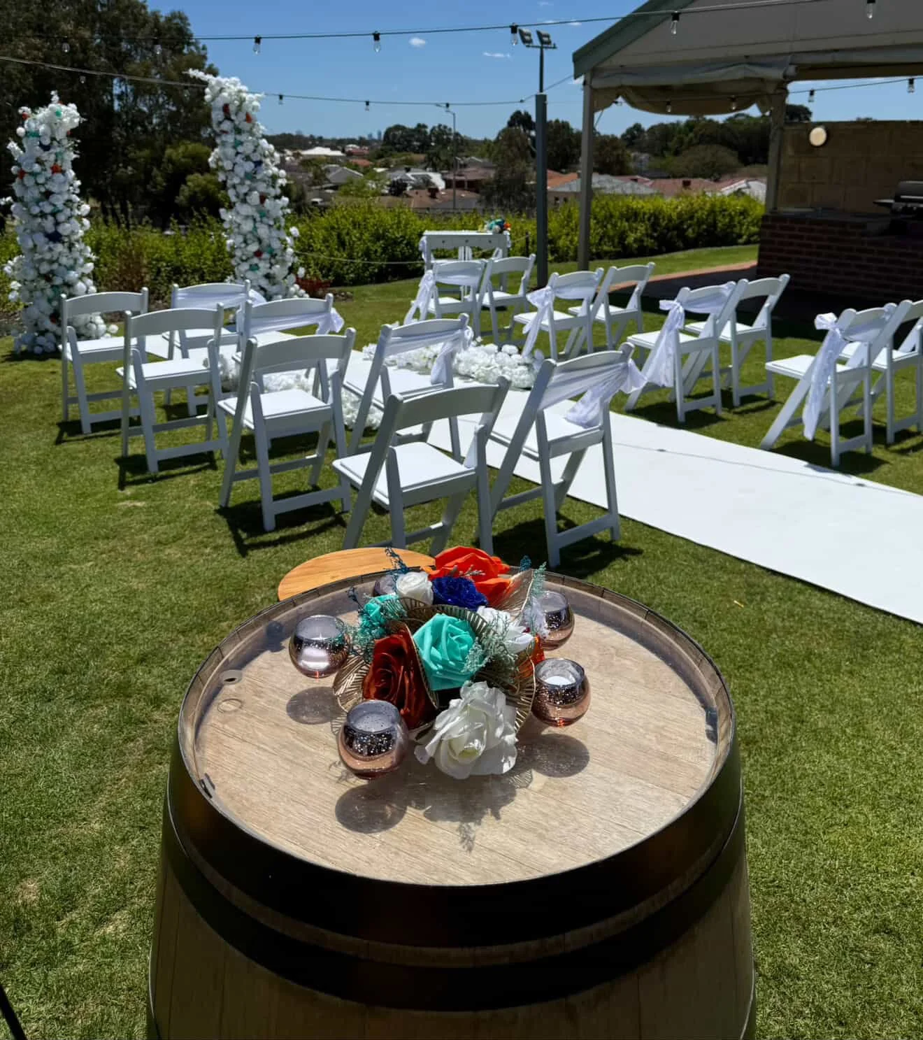 Outdoor wedding setup with white chairs, floral arch, aisle runner, and floral decorations on a barrel table.