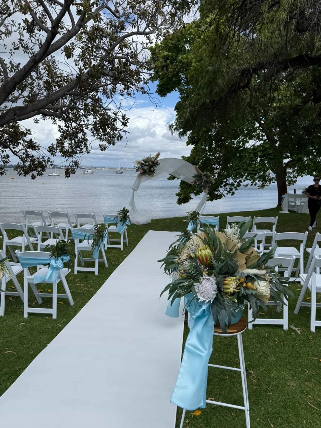 Oceanfront wedding setup with white chairs decorated with blue ribbons, a white aisle runner, floral arrangements with tropical flowers, and a waterfront view with sailboats and cloudy sky
