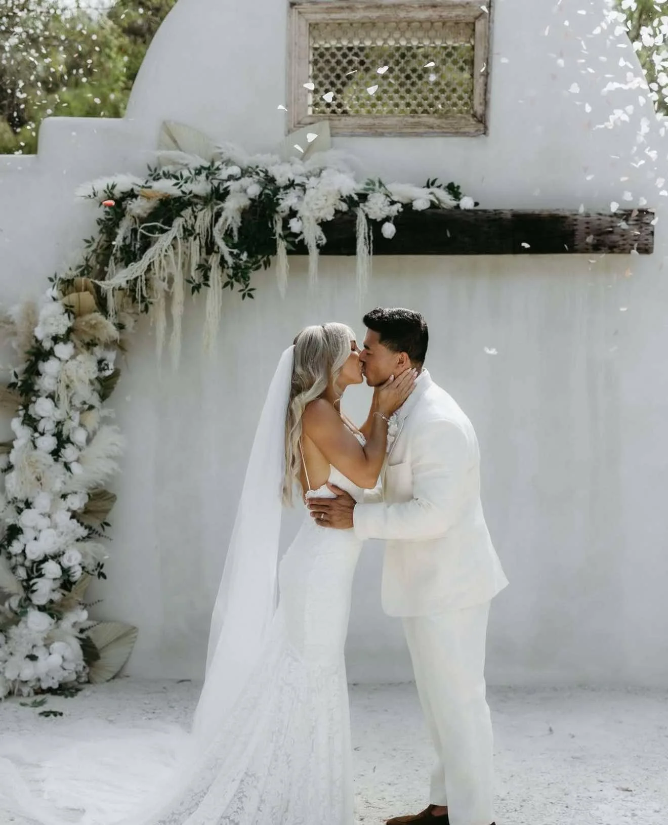 A bride and groom share a kiss during their wedding ceremony outdoors, standing in front of a white wall decorated with flowers and greenery.
