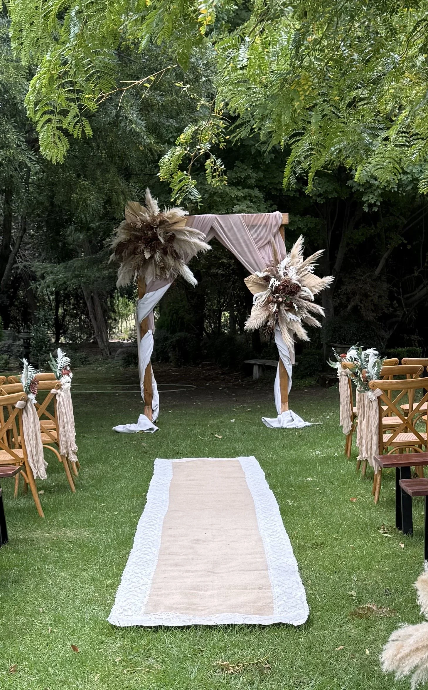 Outdoor wedding ceremony setup with wooden chairs decorated with fabric and flowers, a beige aisle runner on green grass, and a decorative arch made of wood, fabric, and dried flowers and grasses, surrounded by lush green trees.