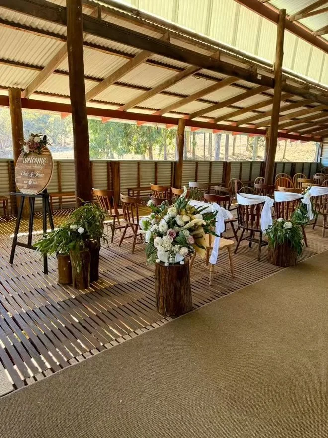 Rustic outdoor event space with wooden chairs, white tablecloths, floral centerpieces, and a welcome sign on an easel under a metal roof.